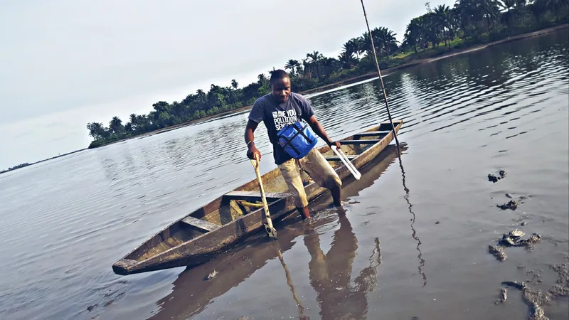 A man stands beside a dugout canoe in the shallow water of Bodo Creek, Rivers State, carrying sampling equipment, with…