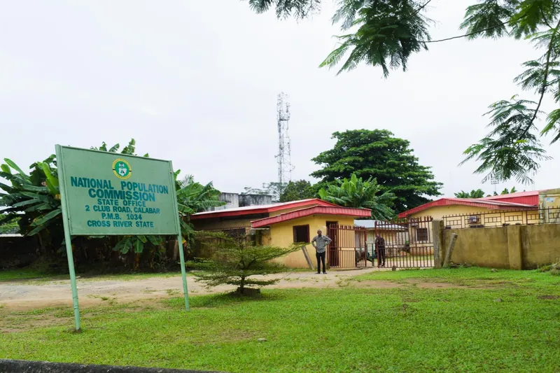 A green signboard reading National Population Commission State Office, 2 Club Road, Calabar, in front of low yellow…