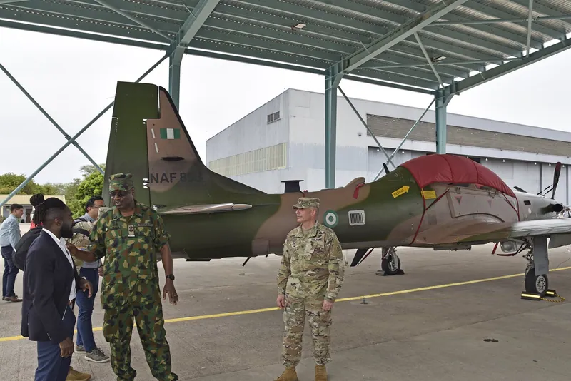 A Nigerian Air Force officer in camouflage talks with visitors beside a green and brown A-29 Super Tucano aircraft…