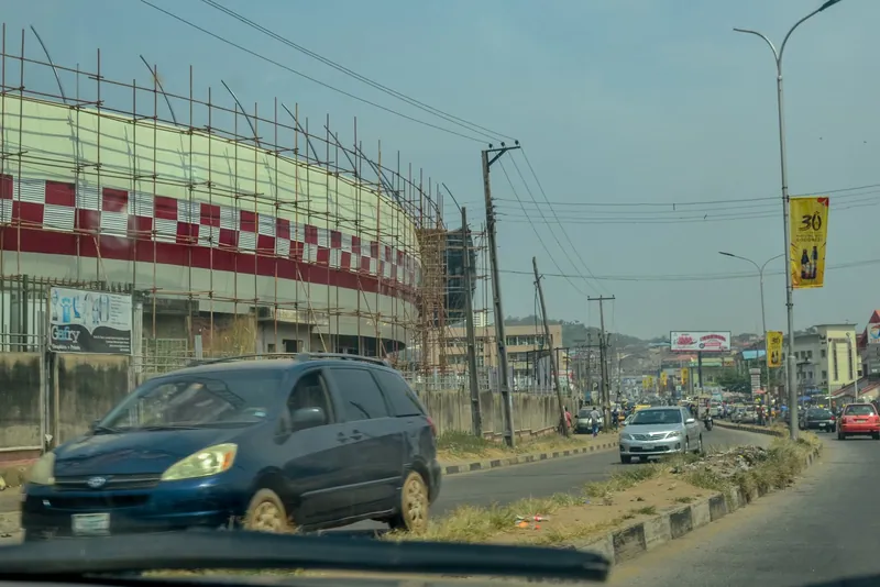 Scaffolding around the curved red and white outer wall of a stadium beside a busy road in Ibadan, seen through a car…