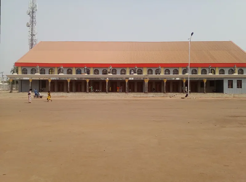 A long two-storey building with an orange roof and a row of statues along its front, behind a wide bare earth square in Lafia