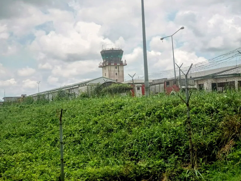 An air traffic control tower rises behind a chain link security fence topped with barbed wire and a bank of green…