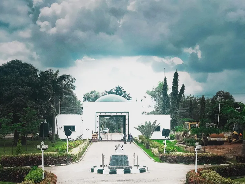 A paved driveway lined with clipped hedges and lamp posts leads to the white domed entrance gate of Kaduna State…