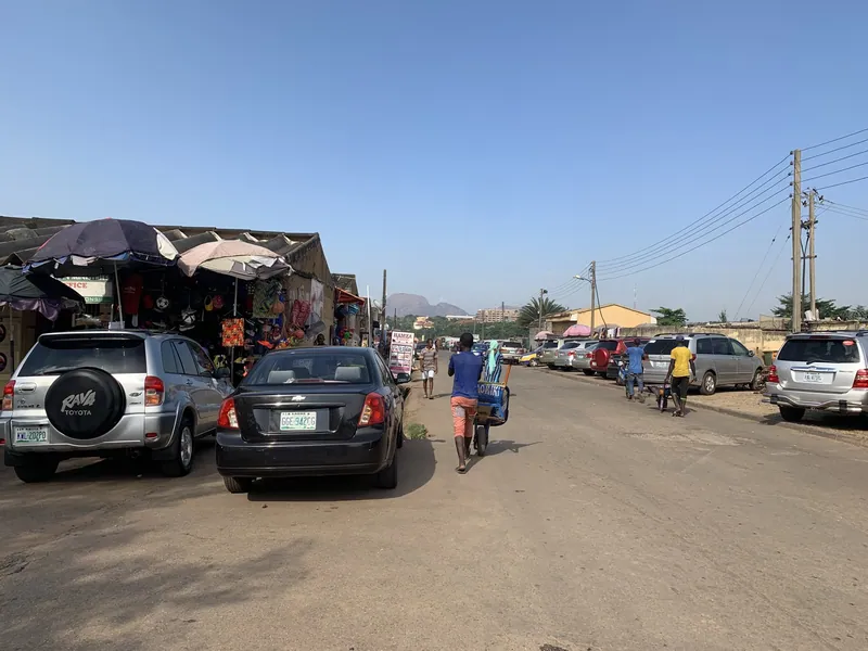 A dusty market road in Garki, Abuja, with umbrella-shaded stalls on the left, parked cars on both sides and a man…