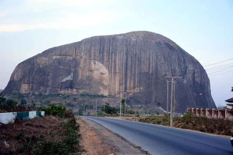 Zuma Rock, a vast grey granite dome streaked with dark stains, rising behind electricity poles and scrub beside a…