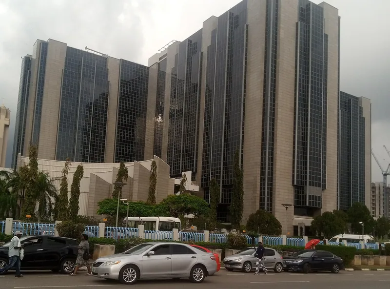 The Central Bank of Nigeria headquarters building in Abuja under an overcast sky, with cars on the road in front