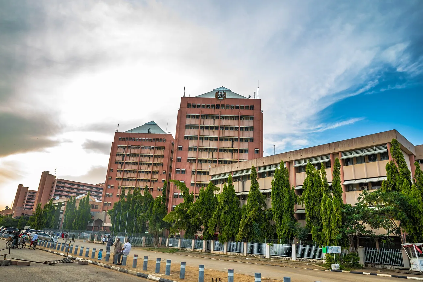 The Federal Secretariat Complex in Abuja, a group of brick-red office towers that house federal ministries, seen from the road under a partly cloudy sky