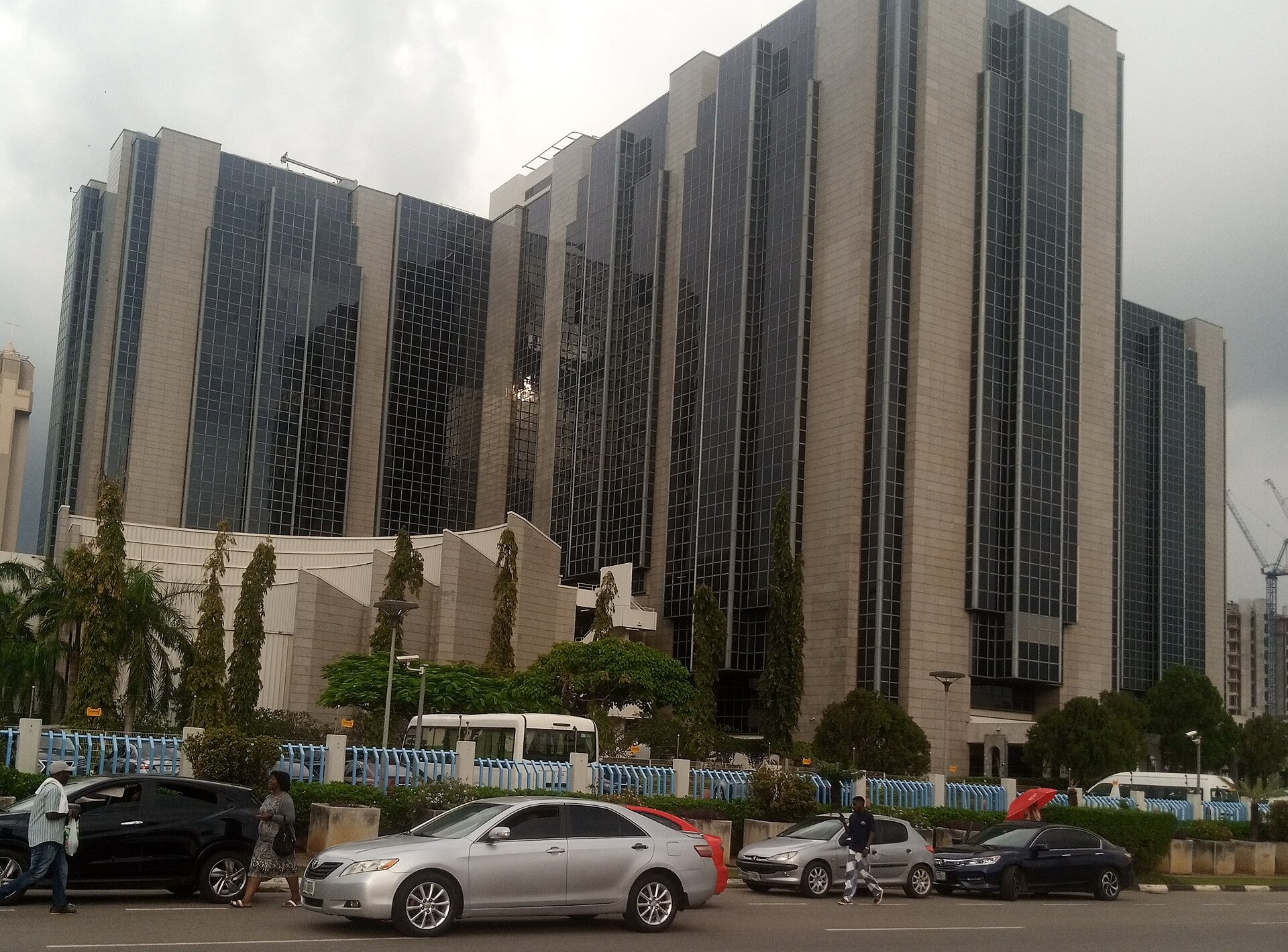 The Central Bank of Nigeria headquarters building in Abuja under an overcast sky, with cars on the road in front